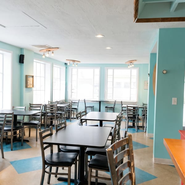 Bright, empty restaurant dining room with blue walls, large windows, and black tables with metal chairs.