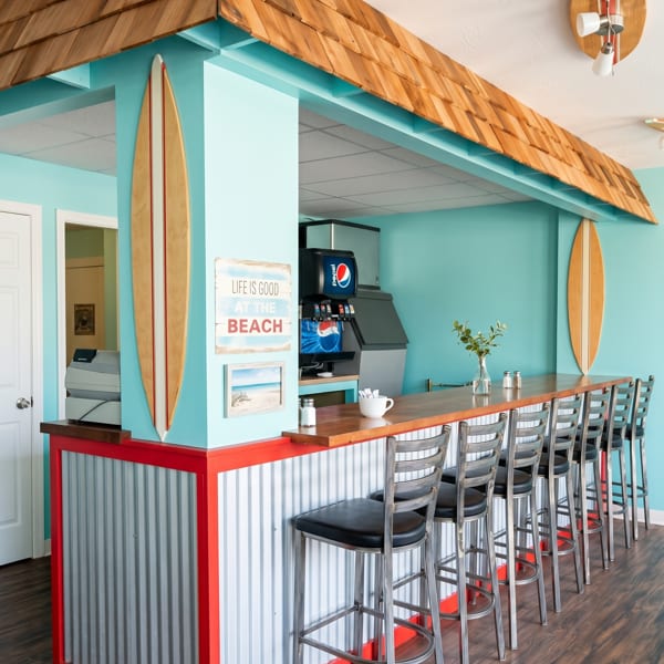 Beach-themed restaurant interior with a long wooden bar, bar stools, and surfboards on turquoise walls.