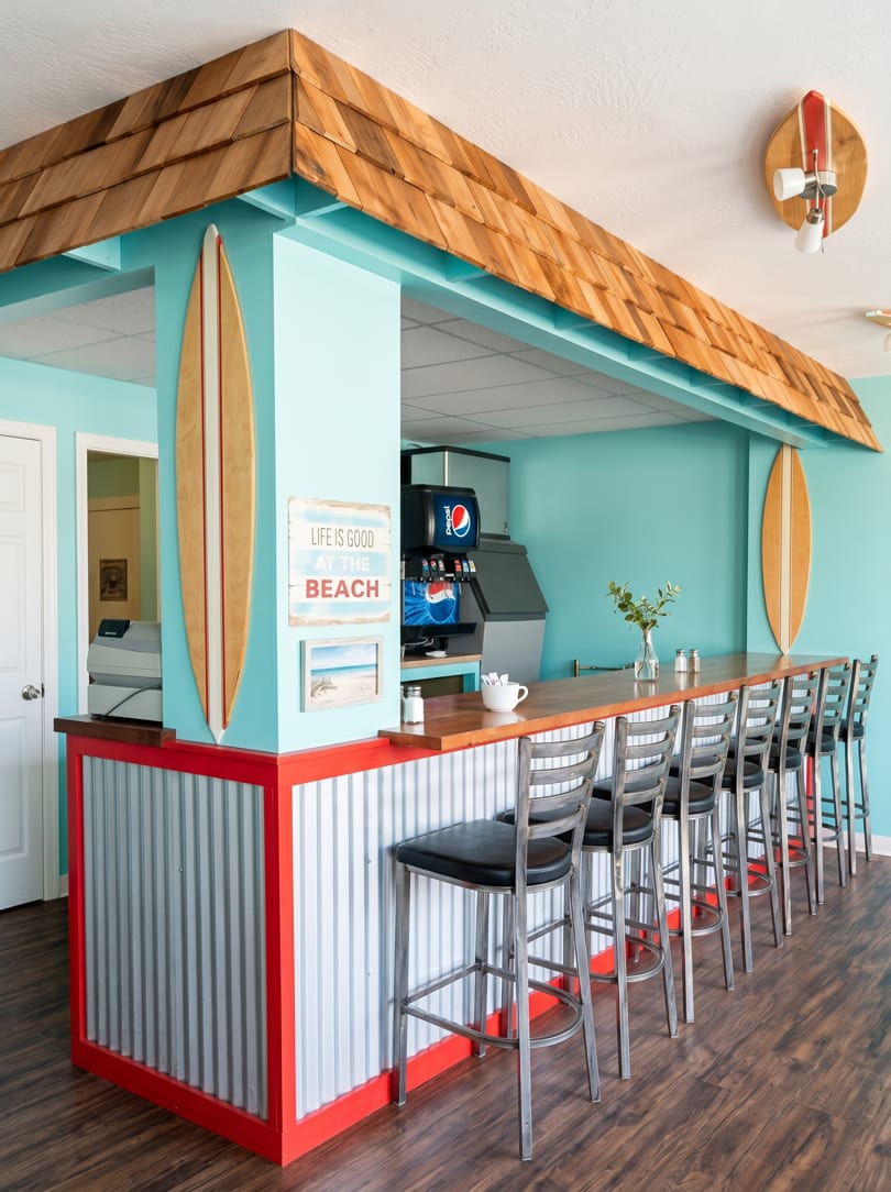 Beach-themed restaurant interior with a long wooden bar, bar stools, and surfboards on turquoise walls.