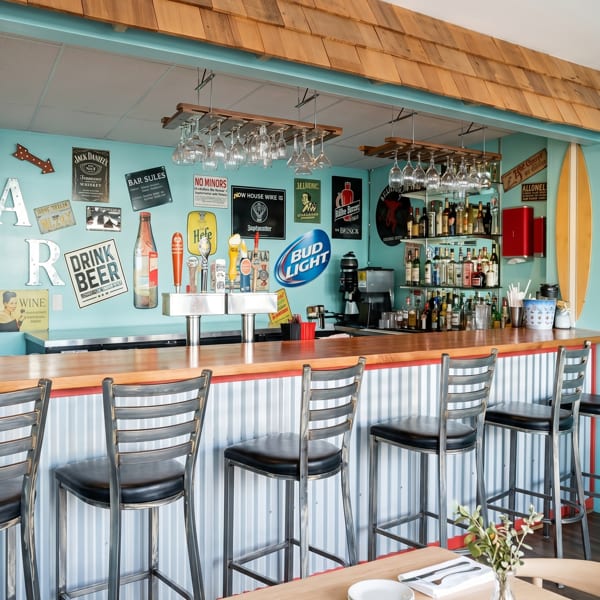 A vibrant bar with a light blue wall, wooden counter, bar stools, and various beer signs.