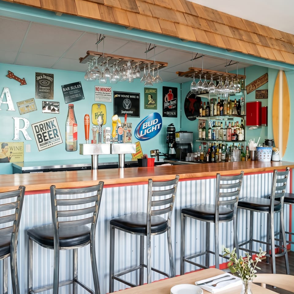 A vibrant bar with a light blue wall, wooden counter, bar stools, and various beer signs.