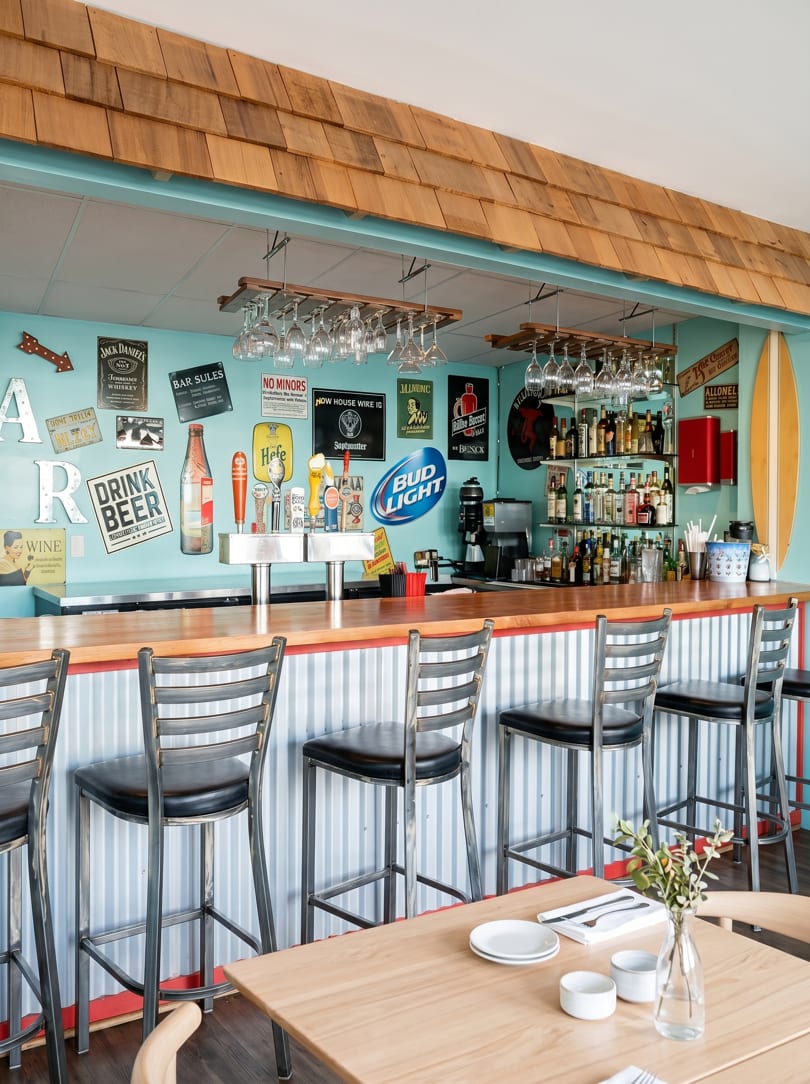 A vibrant bar with a light blue wall, wooden counter, bar stools, and various beer signs.